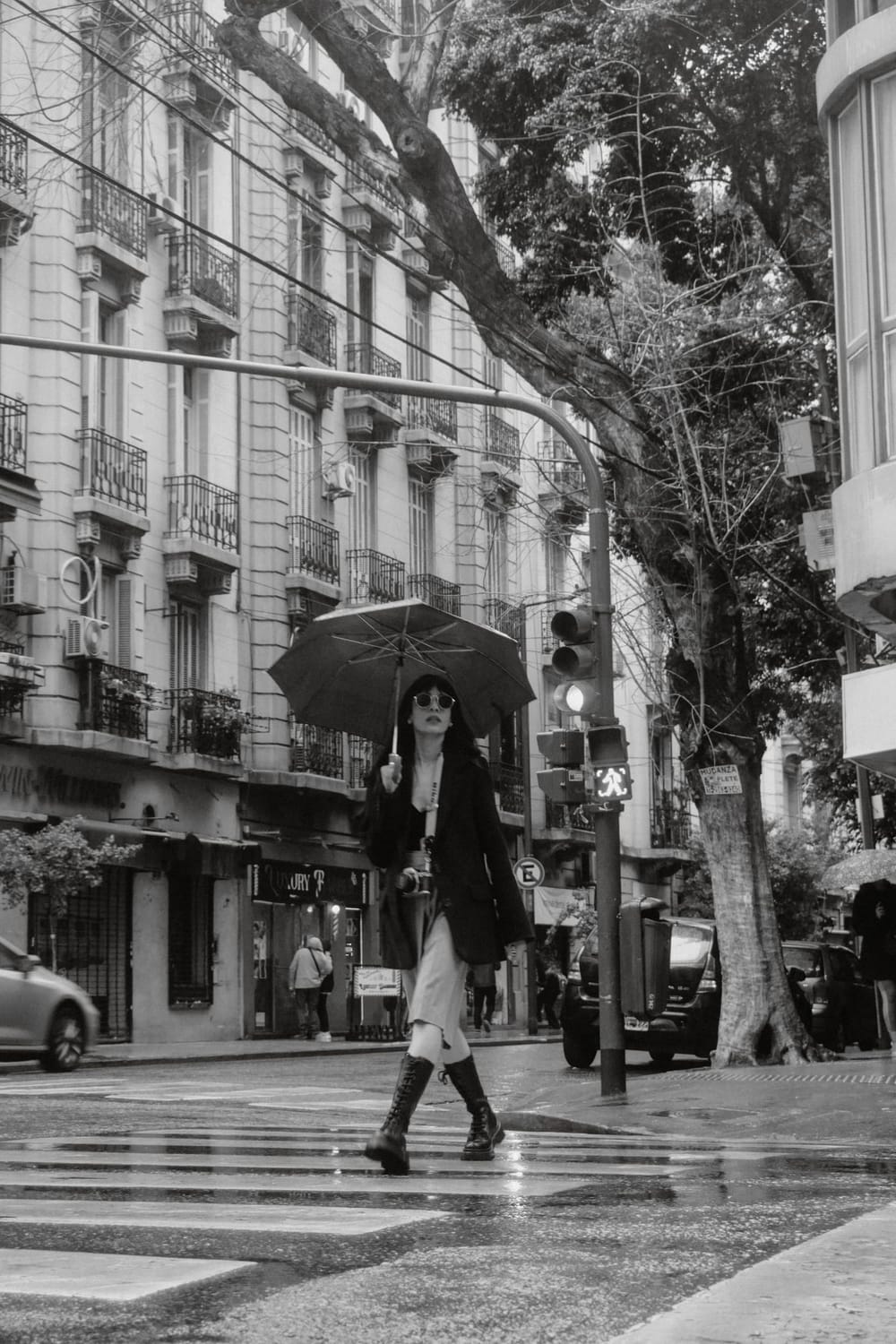 Mujer con paraguas y botas cruzando una calle lluviosa de la ciudad, en blanco y negro